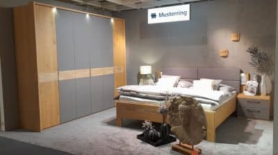 Musterring Schlafzimmer: Holzbett, graue Bettwäsche, passender Kleiderschrank, Dekoartikel auf Teppichboden von Möbel Gruber.