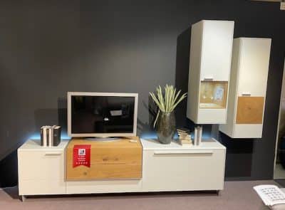 Interliving Wohnwand von Möbel Gruber: Fernseher auf einem schlichten Schrank, Bücher, eine Blumenvase und wandmontierte Aufbewahrungseinheiten.