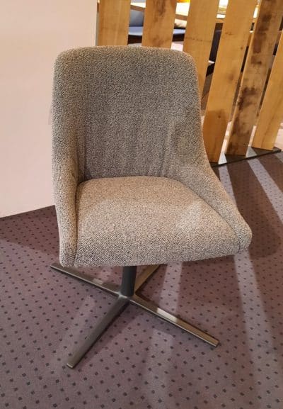 Stuhl von Möbel Gruber: Grau gepolsterter Drehstuhl mit Metallfuß auf gepunktetem Teppich, im Hintergrund Holzpaneele.