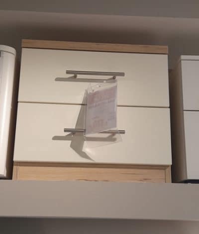 Die Nachtkonsole, ein kleiner Holzschrank mit Metallgriffen, steht auf einem Regal von Möbel Gruber mit einer verschwommenen Beschriftung.