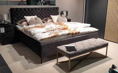 Ein modernes Schlafzimmer von Möbel Gruber verfügt über ein Polsterbett mit Bettkasten, geblümte Bettwäsche, eine Bank und Nachttische mit Lampenaufsatz.