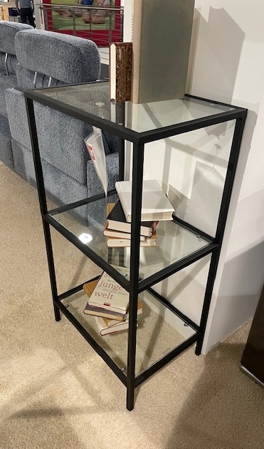 Das Regal von Möbel Gruber, ein schwarzes Bücherregal aus Metall und Glas mit drei Regalböden, steht neben einem gemütlichen blauen Sofa.