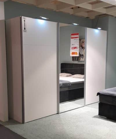 Der Schwebetürenschrank von Möbel Gruber ist ein moderner weißer Schiebetürenschrank mit drei Fächern und Deckenbeleuchtung im Ausstellungsraum.