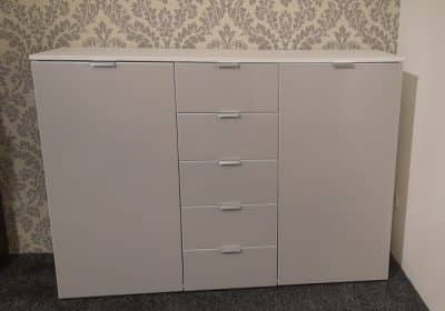 Die moderne weiße Kommode von Möbel Gruber verfügt über fünf Schubladen und zwei Türen, akzentuiert durch eine Tapete mit Blumenmuster.