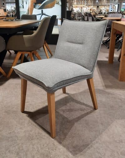 Auf dem gefliesten Boden im Ausstellungsraum von Möbel Gruber steht der elegante Polsterstuhl, ein grauer Stuhl mit Holzbeinen.