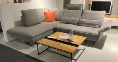Die Polstergarnitur, ein modernes graues Anbausofa von Möbel Gruber mit orangefarbenem Kissen, wird mit einem Couchtisch aus Holz kombiniert.
