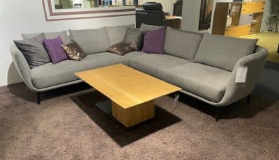Die Schöner Wohnen Polstergarnitur ist ein modernes graues L-Sofa mit lila/braunen Kissen und einem Holztisch auf einem braunen Teppich.