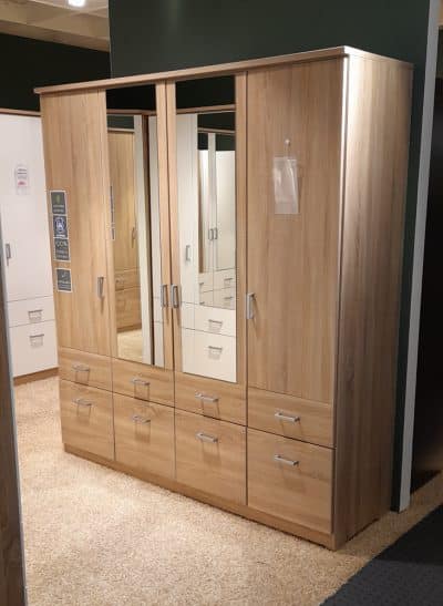 Der Kleiderschrank von Möbel Gruber verfügt über Spiegeltüren und mehrere Schubladen auf einem mit Teppich ausgelegten Ladenboden.