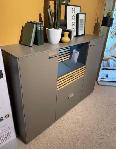Ein modernes graues Interliving-Highboard mit Büchern, einer Pflanze und Dekor darauf vor einer warmen gelben Wand.