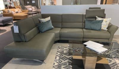 Ein olivfarbenes, modernes Sofa der Polstergarnitur von Interliving mit Kissen steht neben einem Couchtisch aus Glas mit Büchern.