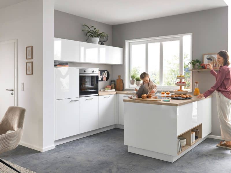 Eine Frau und ein Kind backen gemeinsam in einer hellen, modernen weißen Küche mit großen Fenstern und Grünpflanzen.