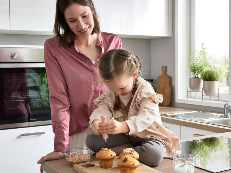 Eine Frau lächelt, während ein junges Mädchen in einer hellen Küche Cupcakes mit Streuseln verziert.