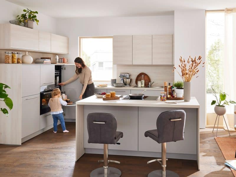 Eine Frau und ein Kind backen gemeinsam in einer modernen, hellen Küche mit hellen Schränken und Pflanzen.